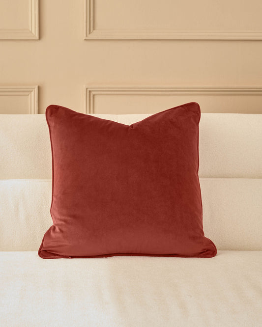 Luca cushion in toffee red velvet displayed on a cream bed against a paneled headboard.