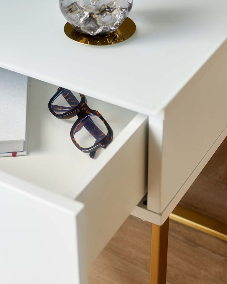 A modern white bedside table with a drawer partly open and gold legs. Glasses and a book are placed in the drawer.