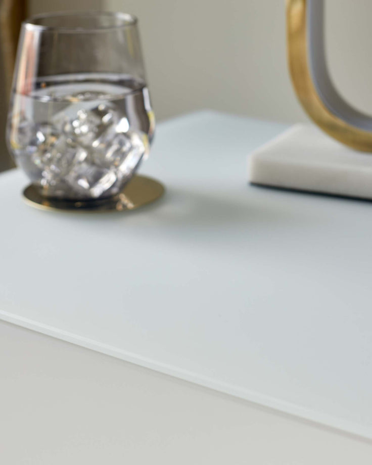 Close up of a glass of water with ice and a gold coaster on a modern white table. Perfect for home offices and bedrooms.