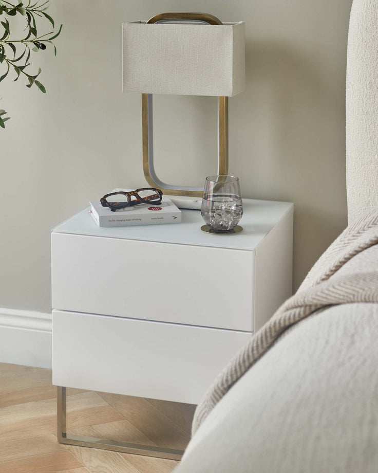 White bedside table with lamp, glasses and book on top. Modern design with metal legs, drawers and glass top.