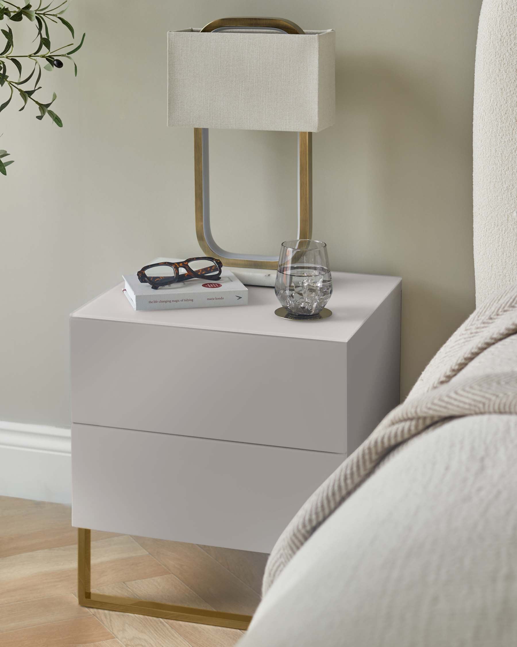 A bedside table with a lamp, book, spectacles and glass of water, next to a bed.