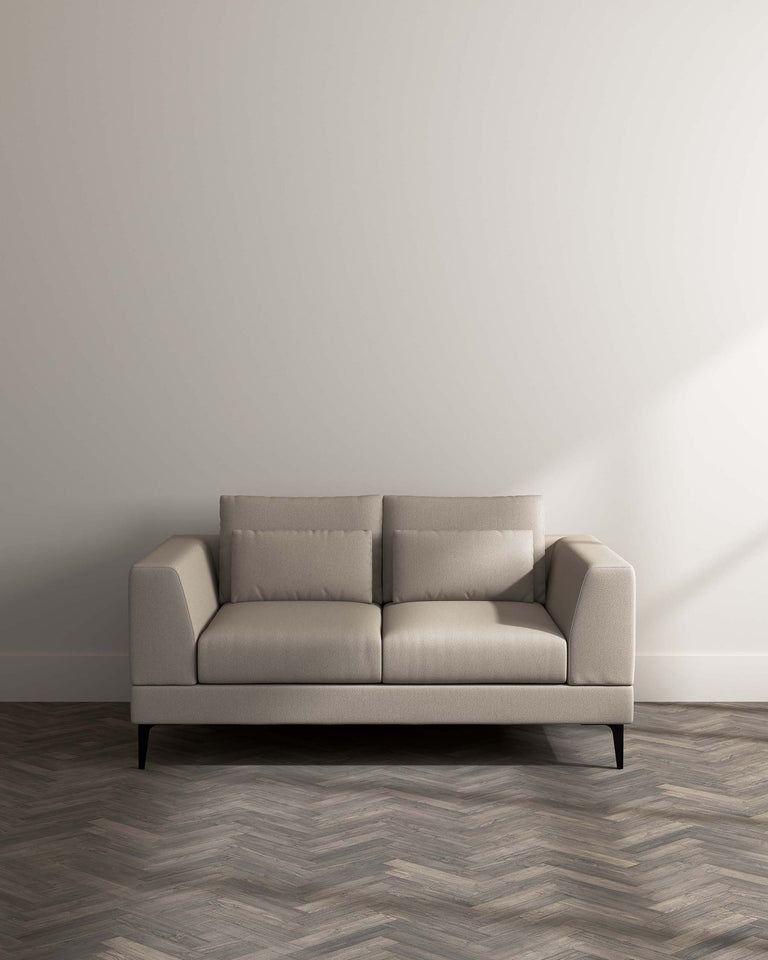 Modern three-seater sofa with a minimalist design, featuring upholstered cushions in a light beige fabric, clean lines, a low-profile backrest, square armrests, and slender black metal legs, set against a neutral backdrop and positioned on a herringbone-patterned wooden floor.