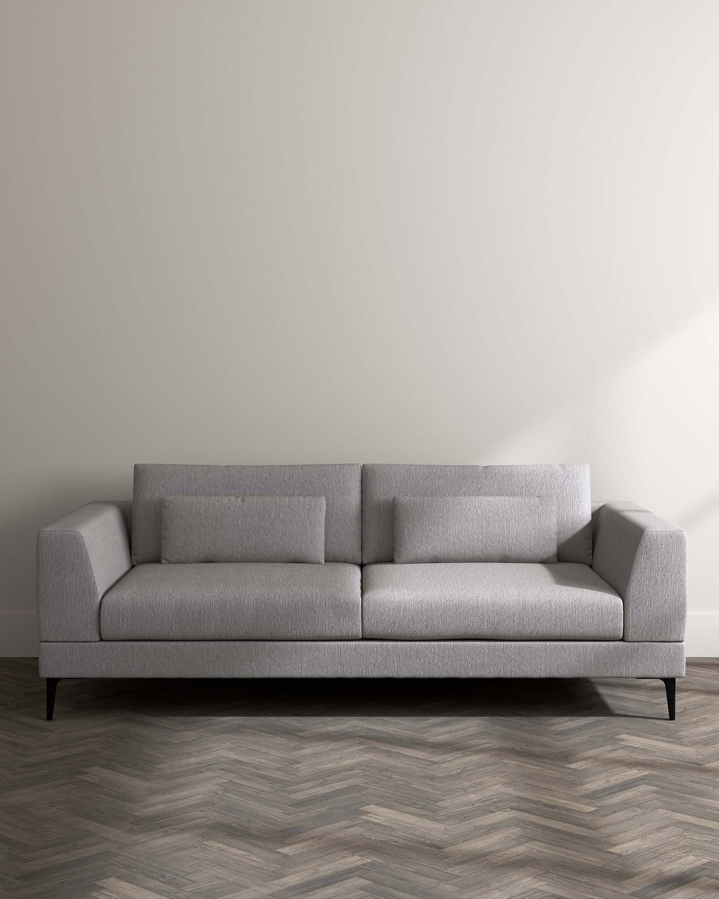 A contemporary light grey fabric sofa with clean lines, featuring plush back cushions, a tufted seat, and angular armrests, raised on slender black metal legs.
