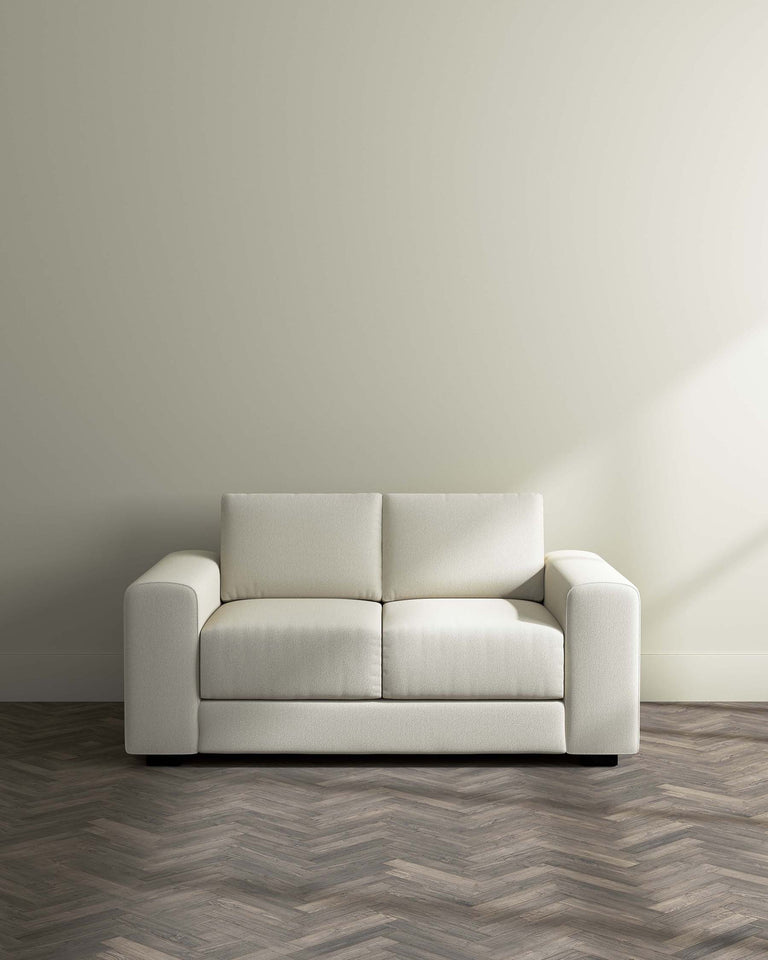 A modern, minimalist off-white three-seat sofa with a solid and structured design, featuring broad armrests and a slightly textured fabric upholstery. The sofa sits on a dark, herringbone-patterned wood floor against a plain, light beige wall with soft lighting.