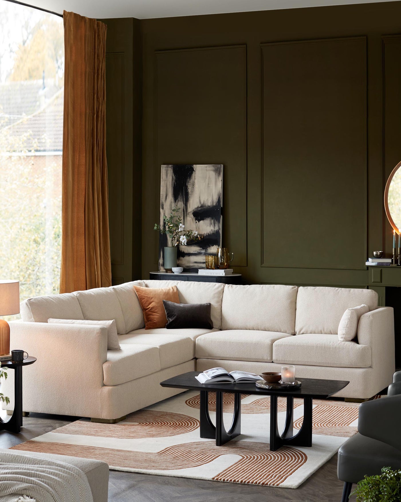 Modern living room setup featuring an L-shaped modular sofa in a light beige fabric with plush cushions, complemented by a set of two nesting coffee tables with black tops and unique curved-base design. A textured off-white and terracotta rug anchors the space.