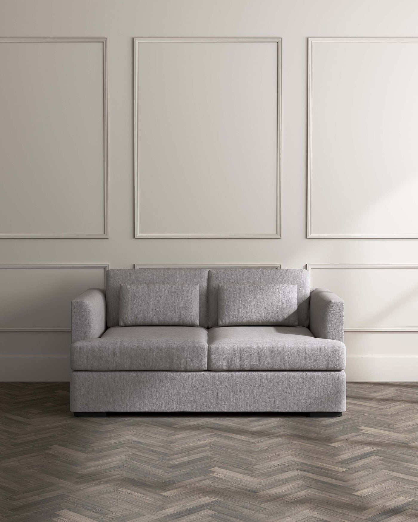 A contemporary grey fabric sofa with clean lines, featuring deep cushioned seats, a low profile, boxy frame, and square armrests, standing on a herringbone patterned wooden floor against a wall with simple framed decorative mouldings.