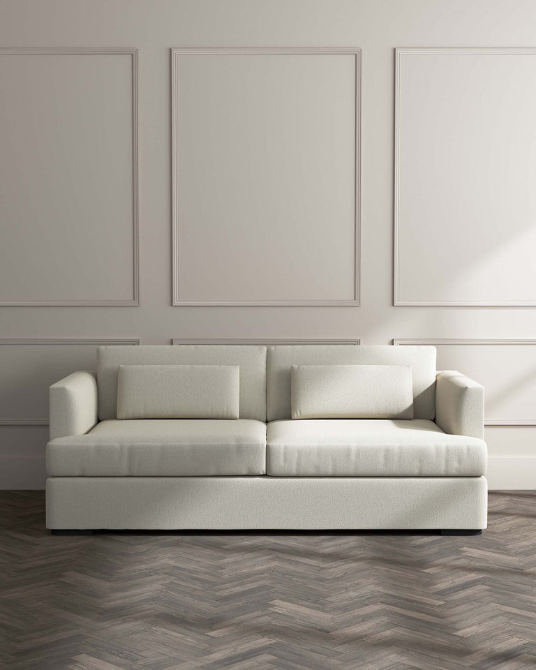 A modern light beige upholstered three-seater sofa with a minimalist design, featuring clean lines, boxy frame, and two matching square back cushions. It's set against a neutral wall with two large blank framed artworks, with herringbone patterned wood flooring.