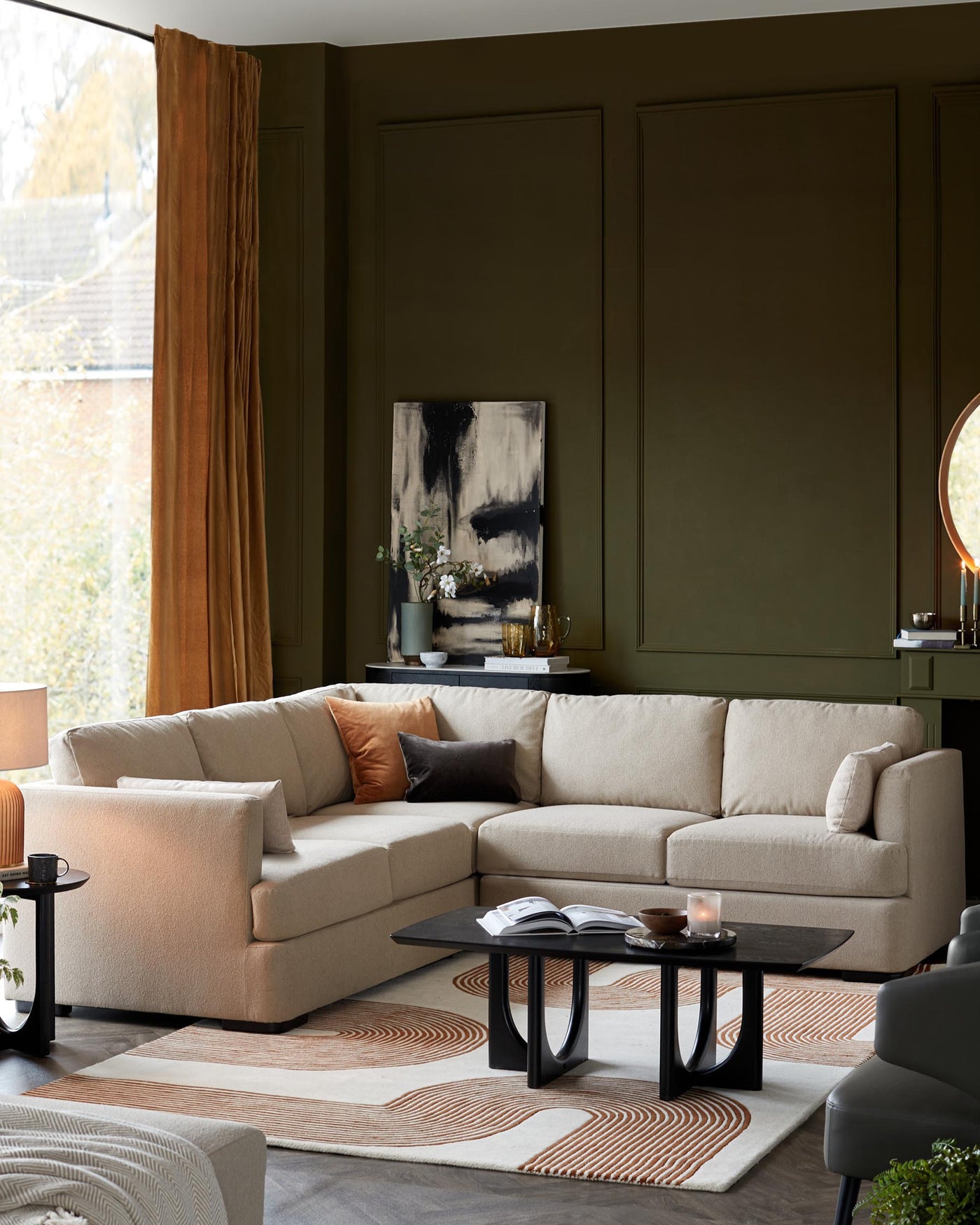 A contemporary modular sofa in a warm beige fabric dominates the space, with plump back cushions and a chaise segment for lounging. A sleek black coffee table with an organic, curved base and a matching round side table contribute minimalist elegance. These pieces are complemented by a soft, geometric-patterned area rug in neutral tones, adding a layer of texture and interest to the room.