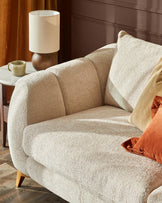 Genevieve 2-seater sofa in natural weave fabric, shown at an angle with curved arms and wooden legs, styled with cushions and lamp.