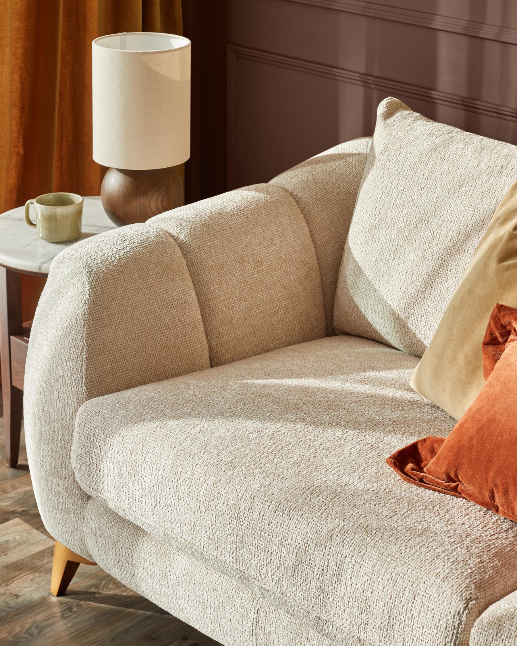 Genevieve 2-seater sofa in natural weave fabric, shown at an angle with curved arms and wooden legs, styled with cushions and lamp.