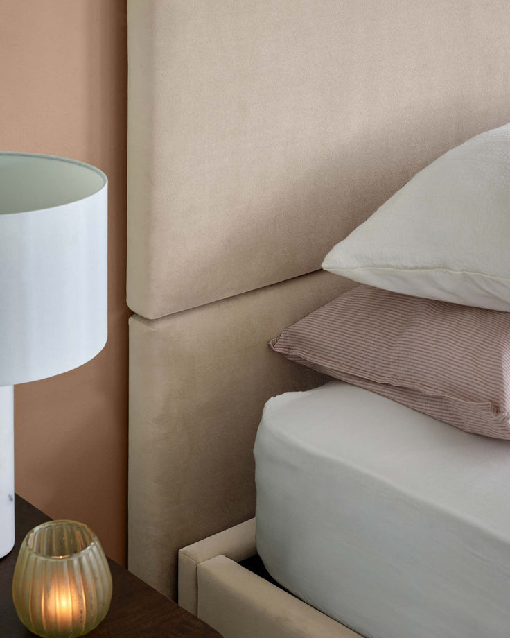 A cozy bed with a soft beige upholstered headboard, layered pillows, a modern lamp, and a decorative candle on the nightstand.