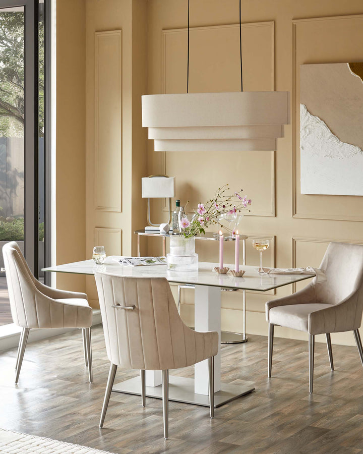 A modern dining set featuring a glass-topped table with four plush, upholstered chairs and a tiered pendant light.