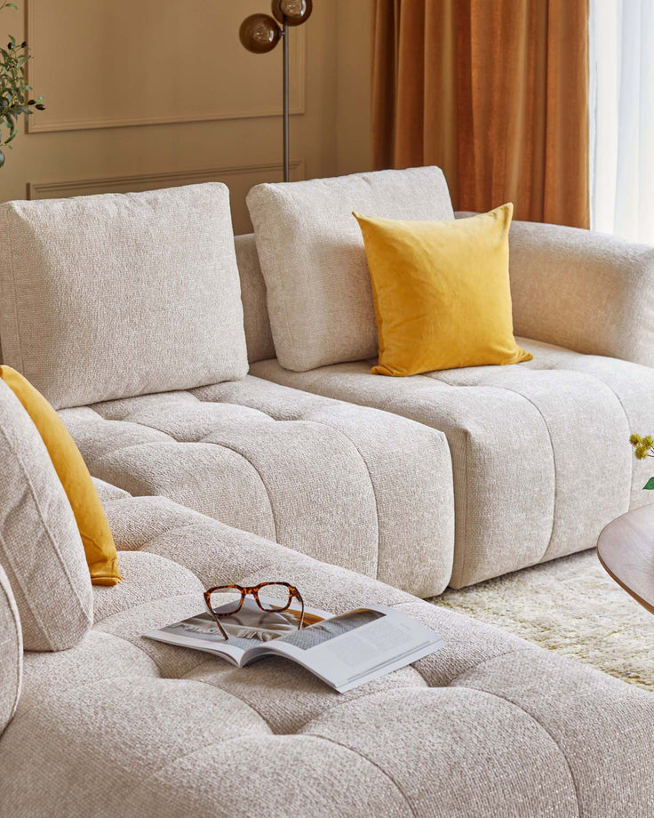 A stylish light beige sectional sofa with textured upholstery, featuring a bright yellow pillow and a coffee table with a magazine and glasses.