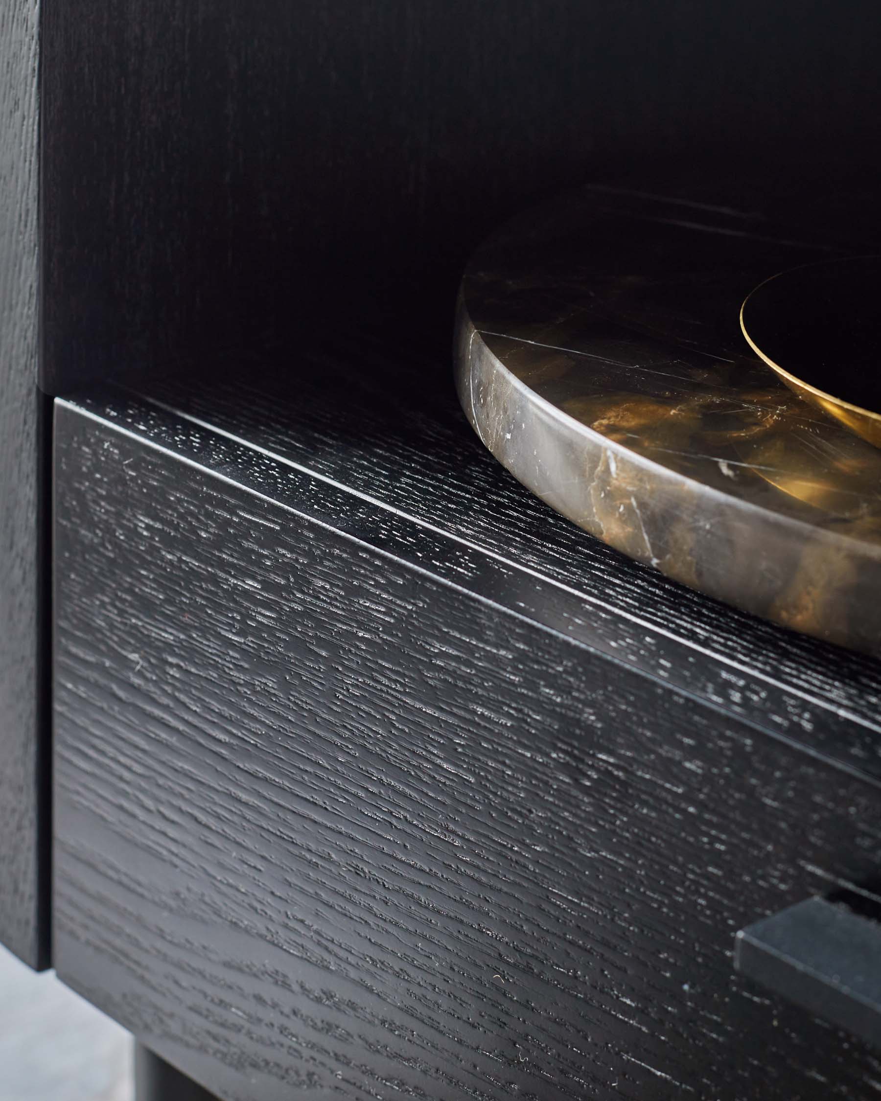 A sleek, dark wood bedside table with a textured finish, topped with a decorative marble and brass bowl.