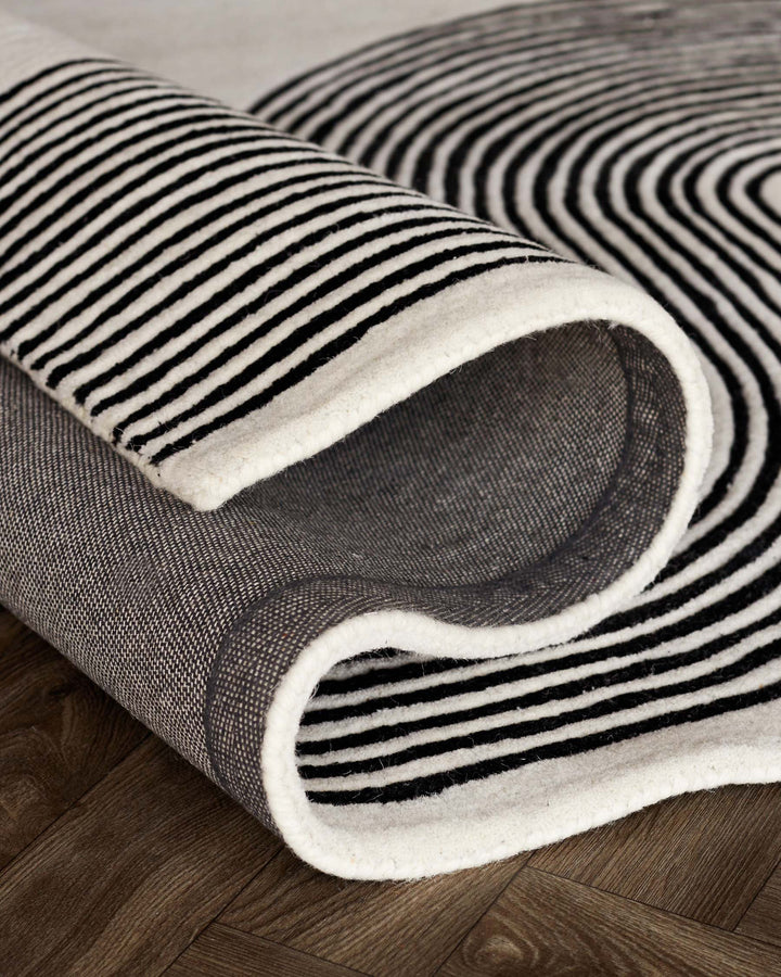 Black and white striped area rug with a modern design, rolled halfway to reveal a textured underside, on a wooden floor.