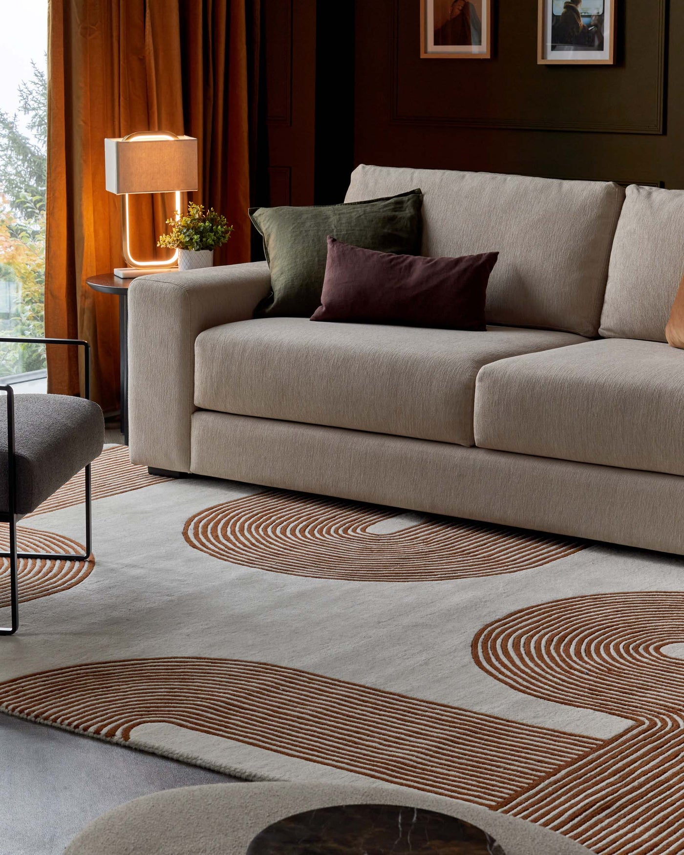 Elegant contemporary beige upholstered three-seat sofa with plush cushions, accompanied by a matching fabric round side chair. A warm copper-toned floor lamp with a geometric design stands atop a sleek circular side table, enhancing the cosy ambiance. The room features a large patterned area rug with swirl motifs in neutral and rust tones, set against a richly textured hardwood floor.