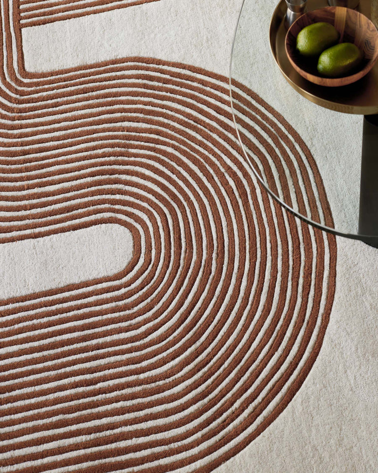 Contemporary area rug with concentric circular patterns in brown and beige, and the edge of a modern round glass top side table with a metallic base, holding a wooden bowl with green limes.