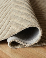 A luxurious beige rug with a subtle chevron pattern, partially rolled on a wooden floor, showcasing its soft texture.