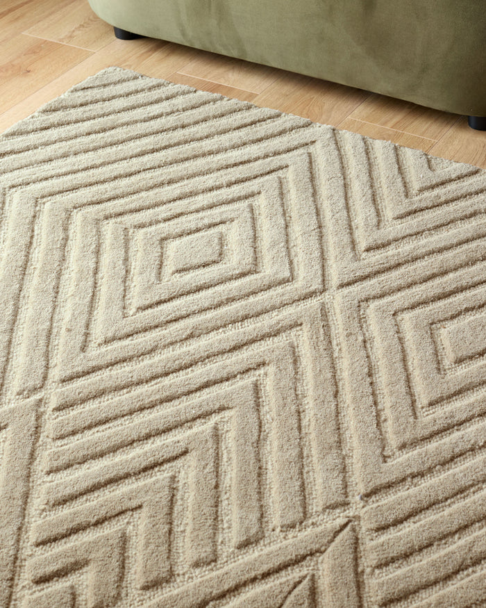 Close up shot of a textured, beige rug with geometric diamond pattern. A small section of a green sofa is also visible.
