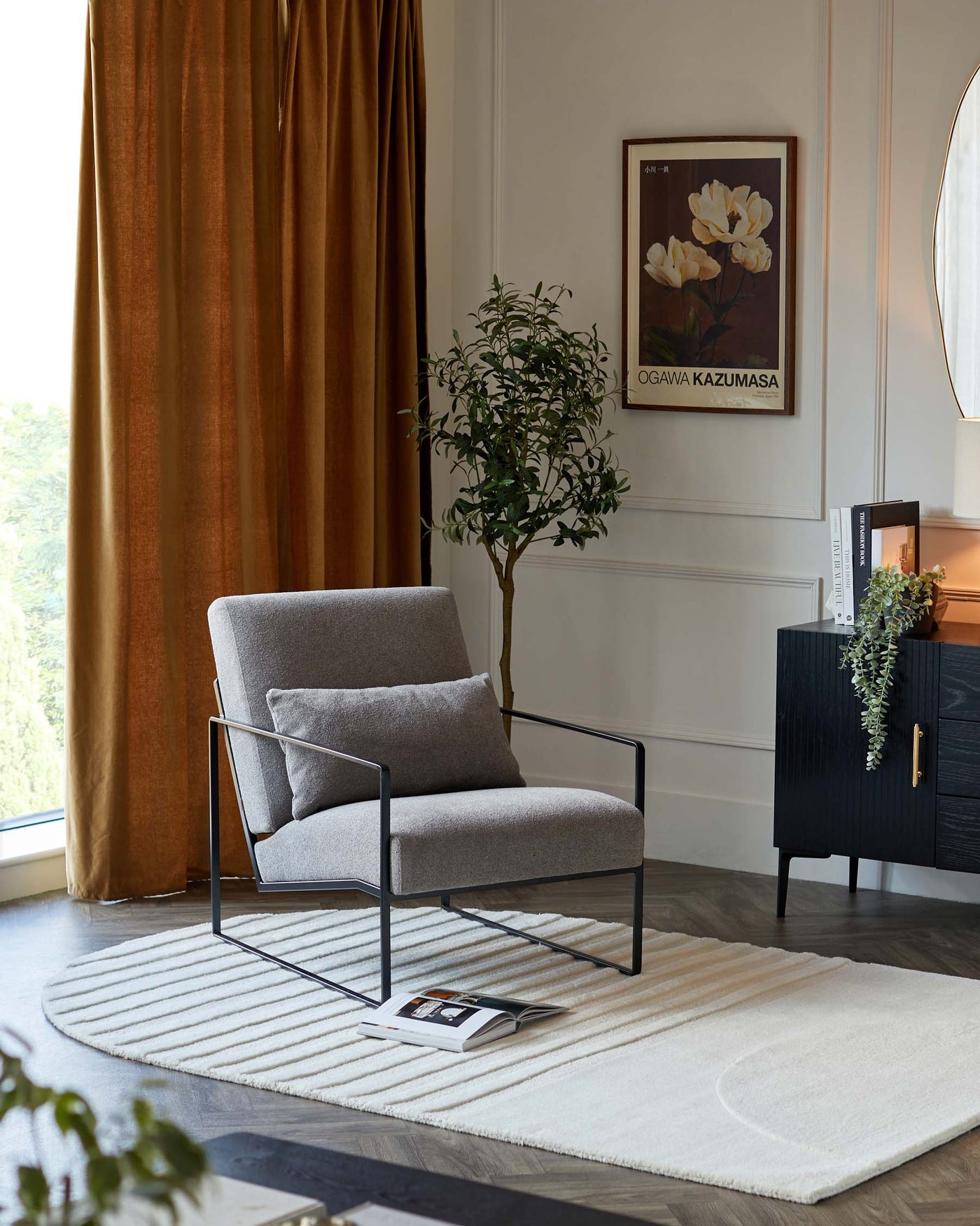 Modern minimalist armchair with a heather-grey upholstery and a slim, black metal frame, beside a small black sideboard cabinet with an understated design and brass hardware accents, all presented on a cream textured area rug.