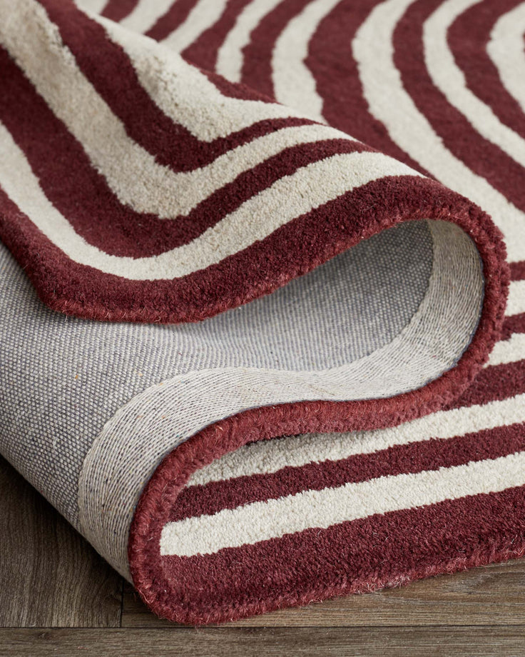 A striped rug with a striking pattern of bold claret and cream-coloured stripes, partially rolled up to reveal its backing.