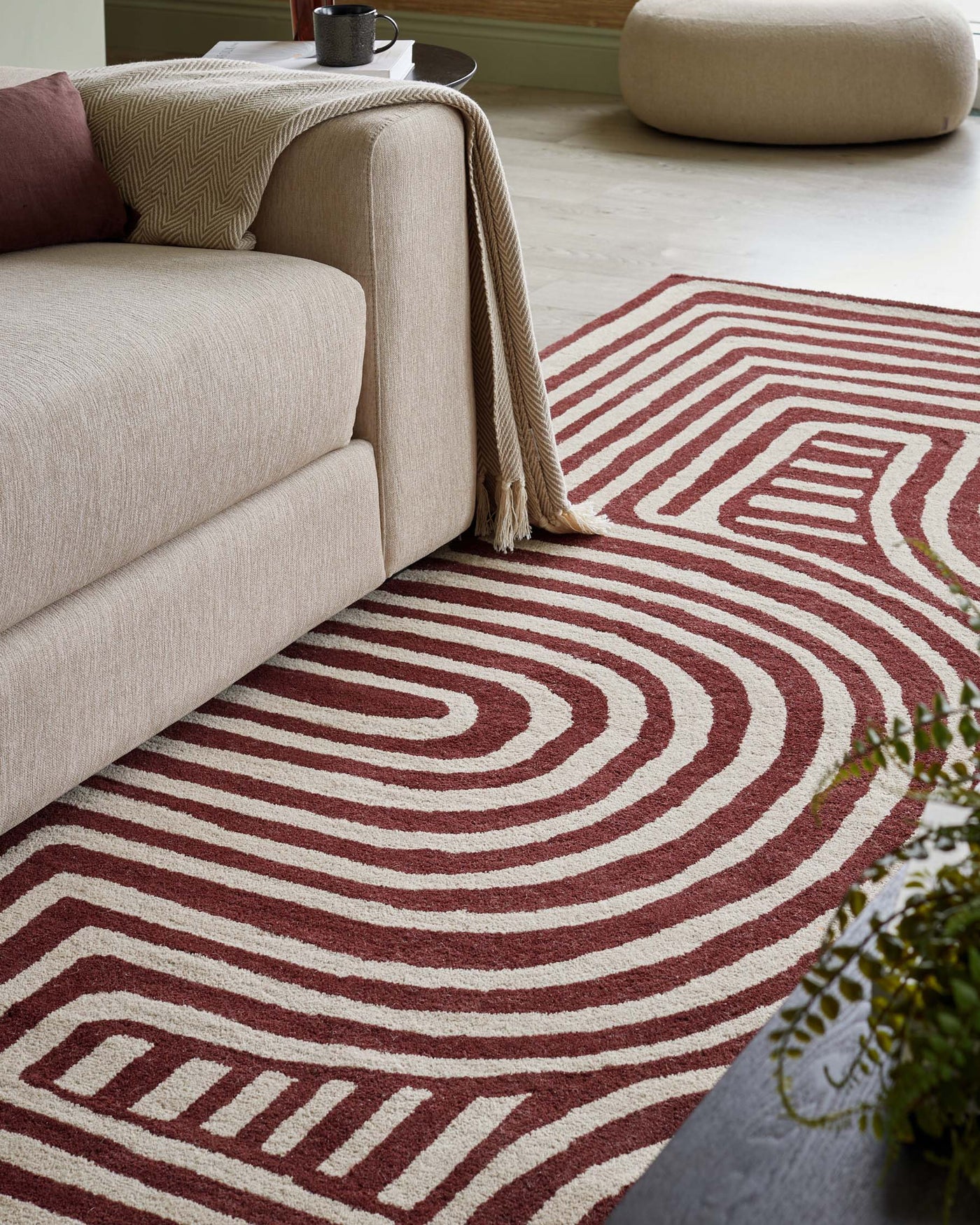 A contemporary taupe sofa with a textured throw blanket is paired with a round side table. In the background, a modern, rounded beige ottoman complements the scene. The geometric patterned area rug beneath features concentric circles in shades of red and cream, creating a bold visual anchor for the space.