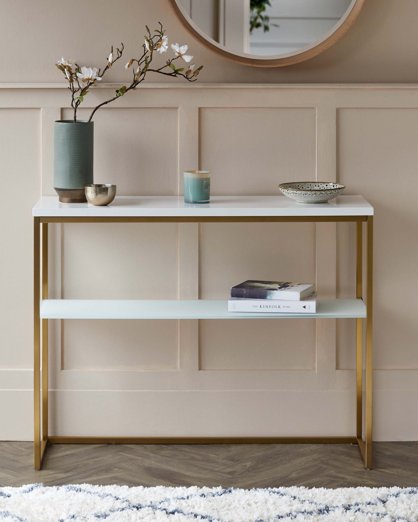 A modern console table with a white rectangular top and a matching lower shelf, supported by sleek brass-finished metal legs, displayed against a panelled wall and above a textured area rug.