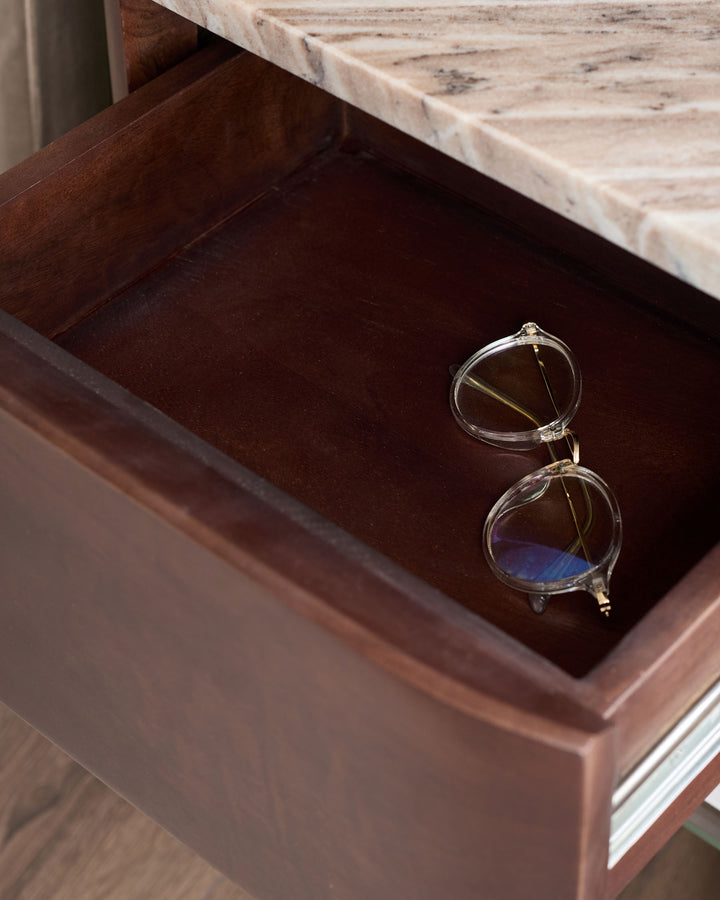 Adalynn bedside table with dark wood drawer open, displaying clear-framed glasses on brown velvet interior, marble top visible.
