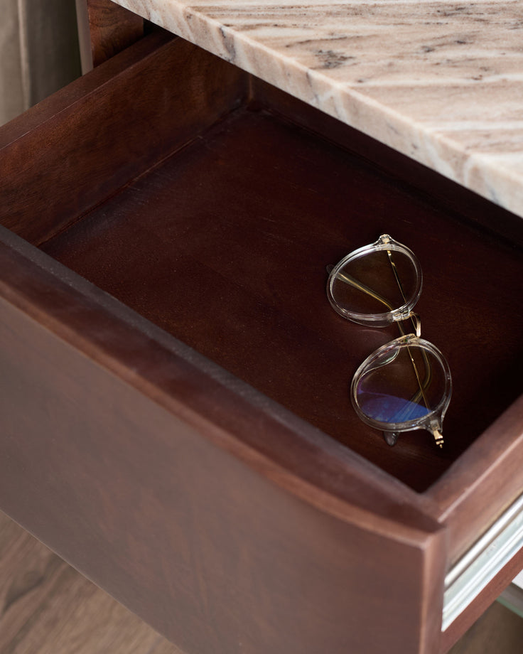 Adalynn bedside table with dark wood drawer open, displaying clear-framed glasses on brown velvet interior, marble top visible.