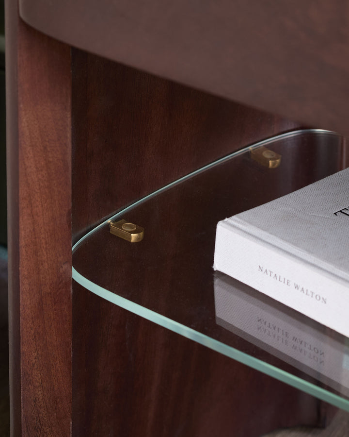 Adalynn bedside table in dark wood with glass shelf, brass hardware, and white book displayed on top.