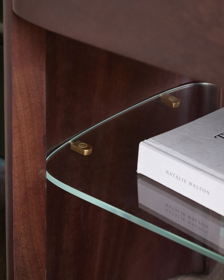 Adalynn bedside table in dark wood with glass shelf, brass hardware, and white book displayed on top.