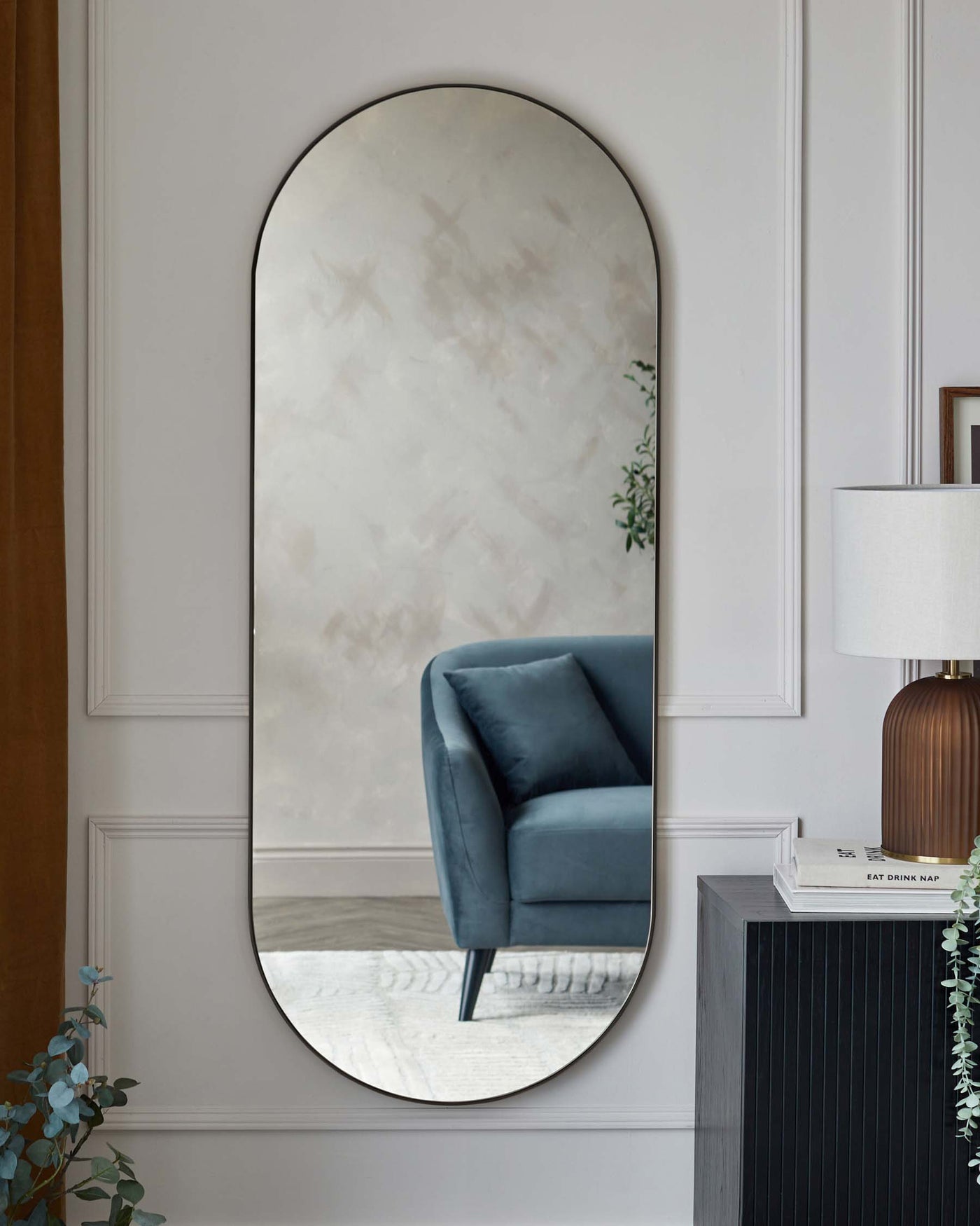 Elegant modern furniture including a plush velvet armchair in a deep blue tone with sleek metal legs, accompanied by a dark-toned wooden sideboard with vertical line detailing, and a classic white lamp with a cylindrical shade and a wooden base. A large, vertical, oval-shaped mirror with a simple frame is also featured, enhancing the room's spacious and sophisticated ambiance.