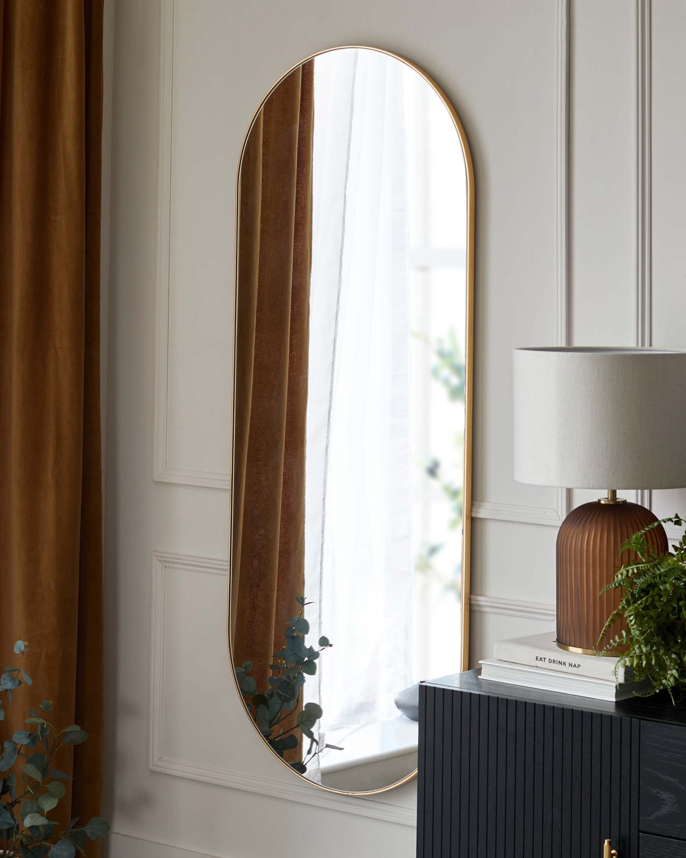 A tall, narrow, wall-mounted arched mirror with a slim gold frame. A black cabinet with vertical grooves and gold knobs. On top of the cabinet is a white lamp with a fluted terracotta base and a white shade, alongside stacked books and a potted green plant.