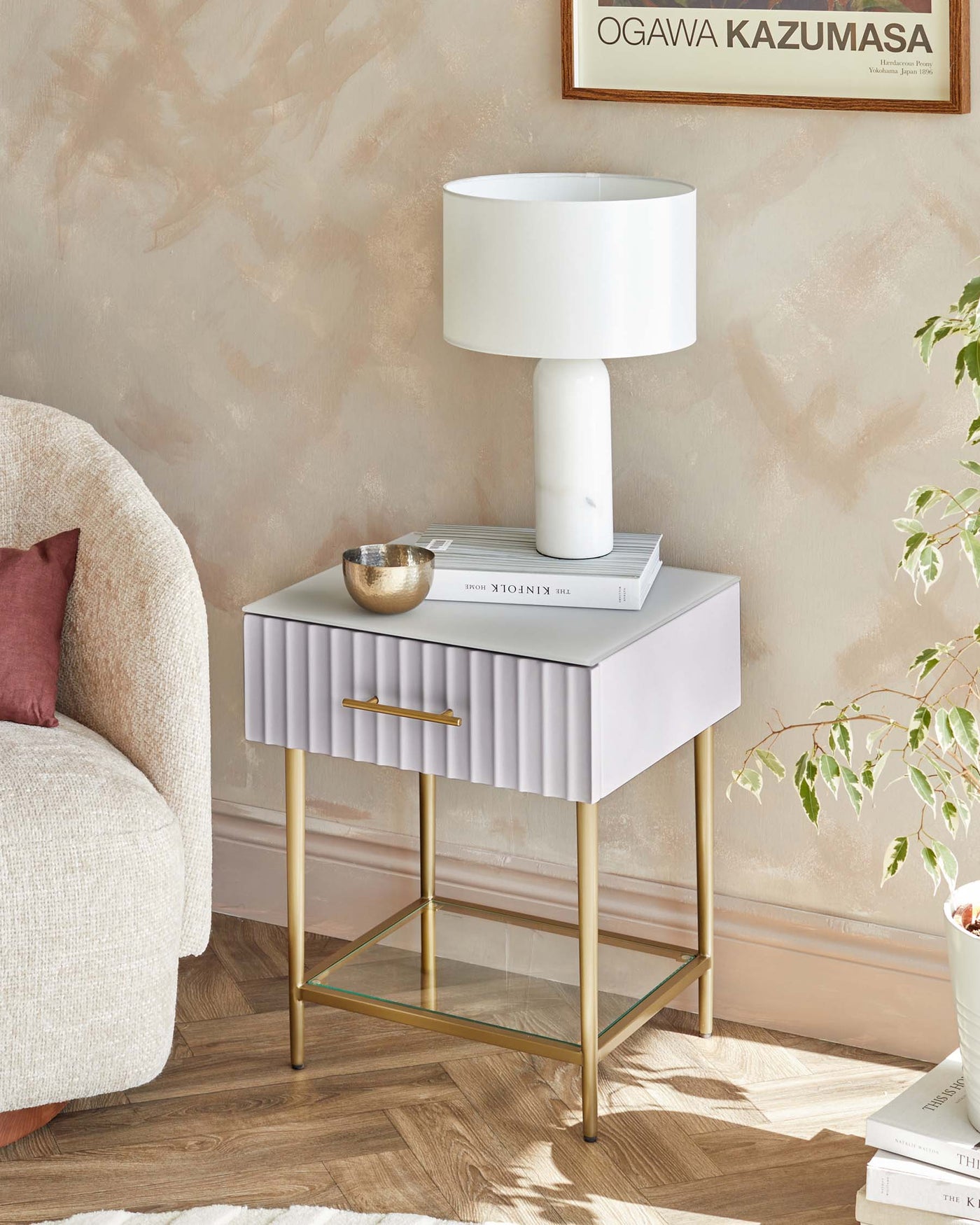 Elegant contemporary side table with a textured, pleated white drawer front, complemented by a sleek brass handle and slender brass legs featuring a lower glass shelf. A chic white lamp and decorative accessories rest atop, enhancing the modern aesthetic.