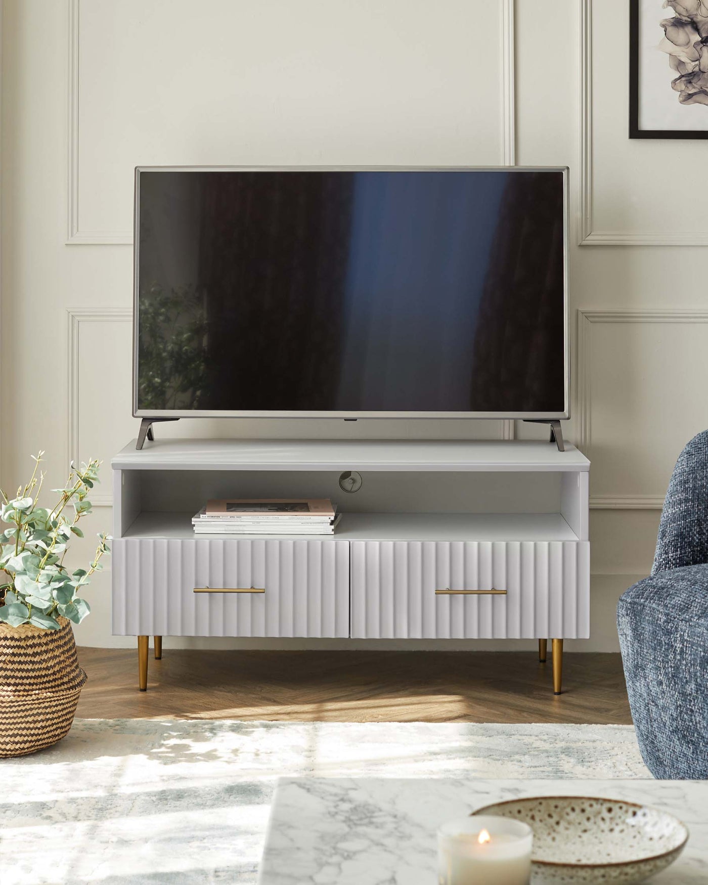 Modern light grey media console with textured front panels and gold-finished legs, supporting a flat-screen TV. Adjacent to the console is a wicker planter with greenery and a fragment of a blue upholstered chair visible to the right. A decorative marble-patterned rug is partially shown in the foreground.