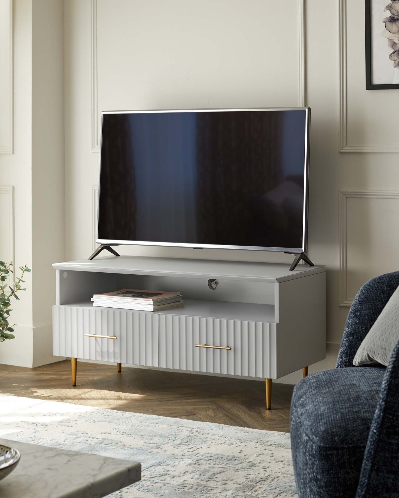 Modern light grey media console with ribbed front drawer detail and brass finished elongated handles, supported by gently tapered legs with gold-tone tips. It is positioned against a wall in a well-lit room, topped with a large flat-screen TV, and flanked by a small stack of books to the side.