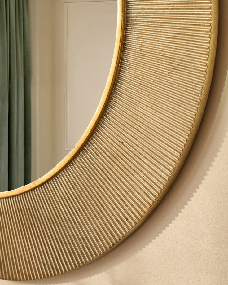 Gold round wall mirror with ribbed texture detail and curved gold frame, mounted on beige wall.