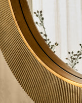 Round gold wall mirror with curved wooden frame, shown at an angle with dried botanicals visible in the background.