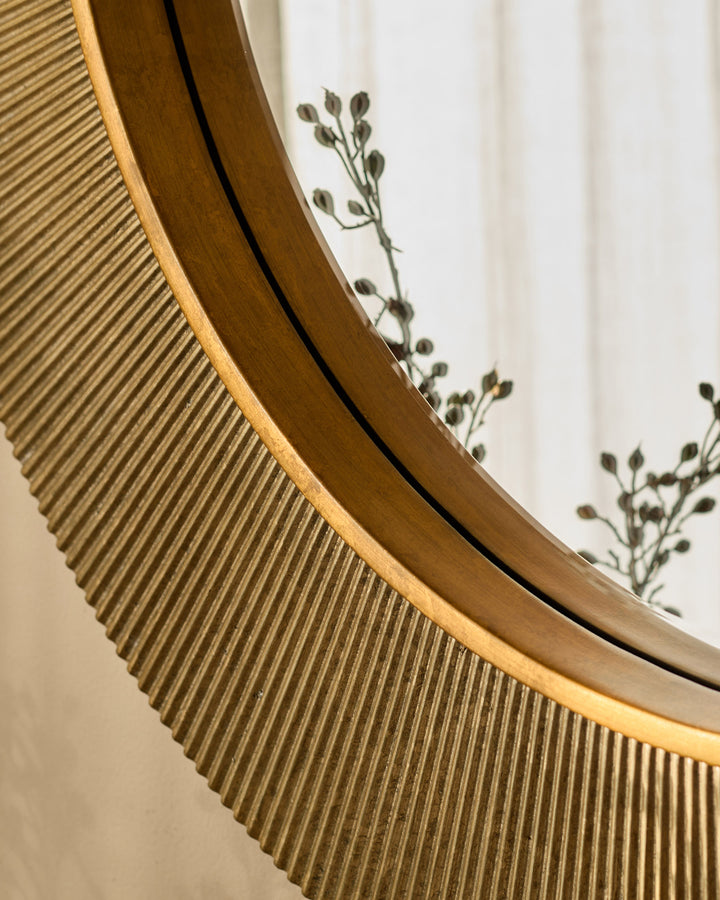 Round gold wall mirror with curved wooden frame, shown at an angle with dried botanicals visible in the background.