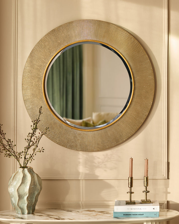 Round gold wall mirror with textured radiating pattern frame, displayed above marble console with decorative accessories.