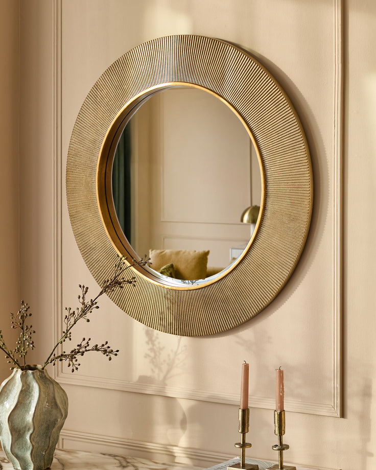 Gold round wall mirror with textured radial pattern frame, displayed above marble surface with candles and dried flowers.