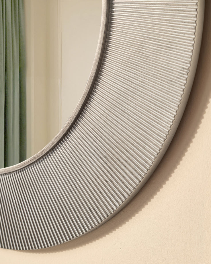 Round silver wall mirror with ribbed texture detail and curved frame, shown at angle against beige wall.