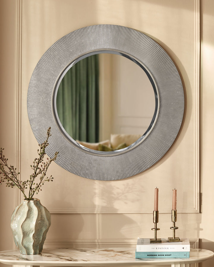 Round silver wall mirror with textured radial line pattern frame, displayed above marble console with vase and candles.