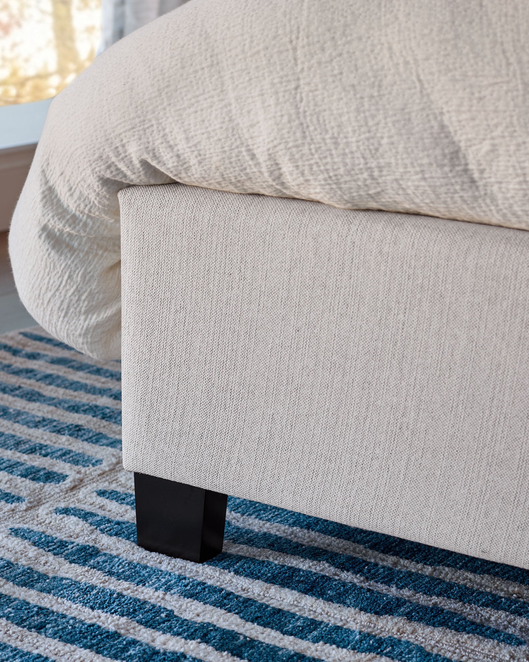 Amalfi fabric bed in ivory with black wooden legs, shown from side angle on striped blue and beige rug.