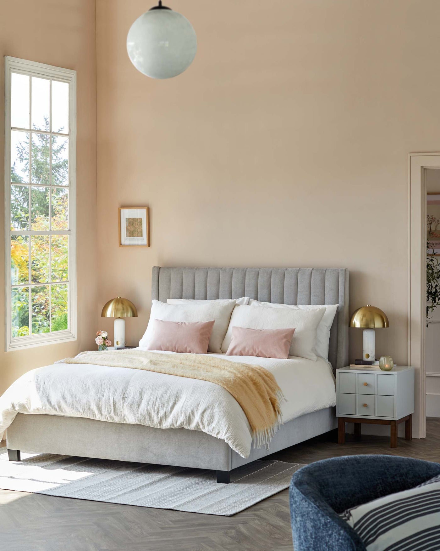 Elegant bedroom setup featuring a grey upholstered king-sized bed with a tufted headboard, accented with white and pale pink bedding and a plush beige throw. Beside the bed is a white two-drawer nightstand with wooden legs, topped with a golden lamp. A striped grey and white area rug lies partially under the bed, complementing a navy blue upholstered chair with white piping in the foreground.