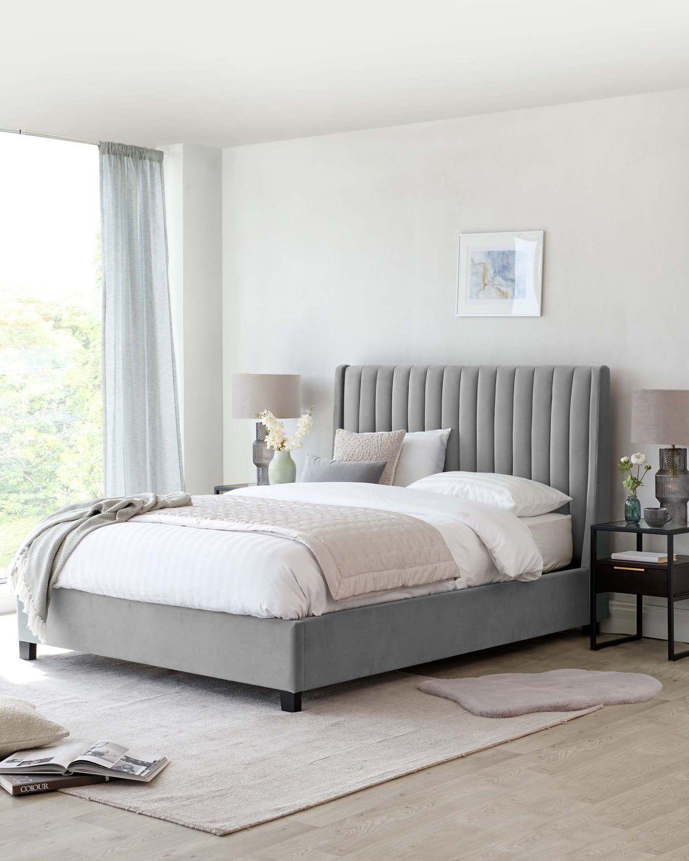 A contemporary-styled bedroom featuring a large upholstered bed with a tall, vertical channel-tufted headboard in a soft grey fabric. The bed is dressed in white linens with a textured comforter and multiple pillows varying in size and shades of grey and white. Beside the bed, there's a small dark wood nightstand with a drawer, accompanied by a simple modern table lamp with a grey shade. The room is carpeted with a large, light beige area rug, complementing the neutral, serene colour palette of the space.