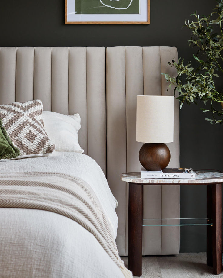 Bedroom scene with a neutral headboard, bedside table with lamp and bed with white duvet and patterned cushions.