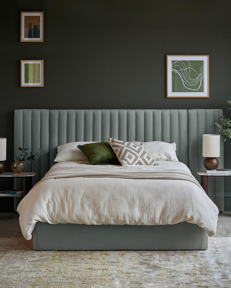A styled bedroom featuring a bed with a padded headboard, side tables and framed artwork on a dark green wall.