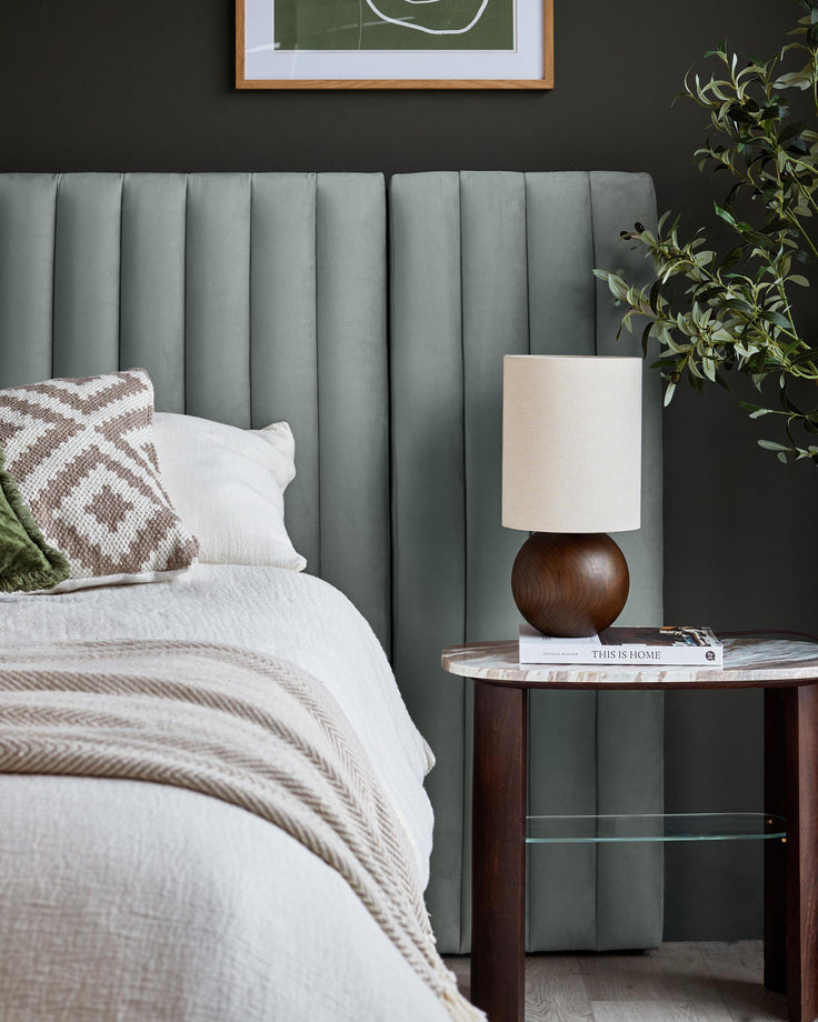 Bed with headboard, side table with lamp, cushion and throw, plants and artwork in a styled bedroom.