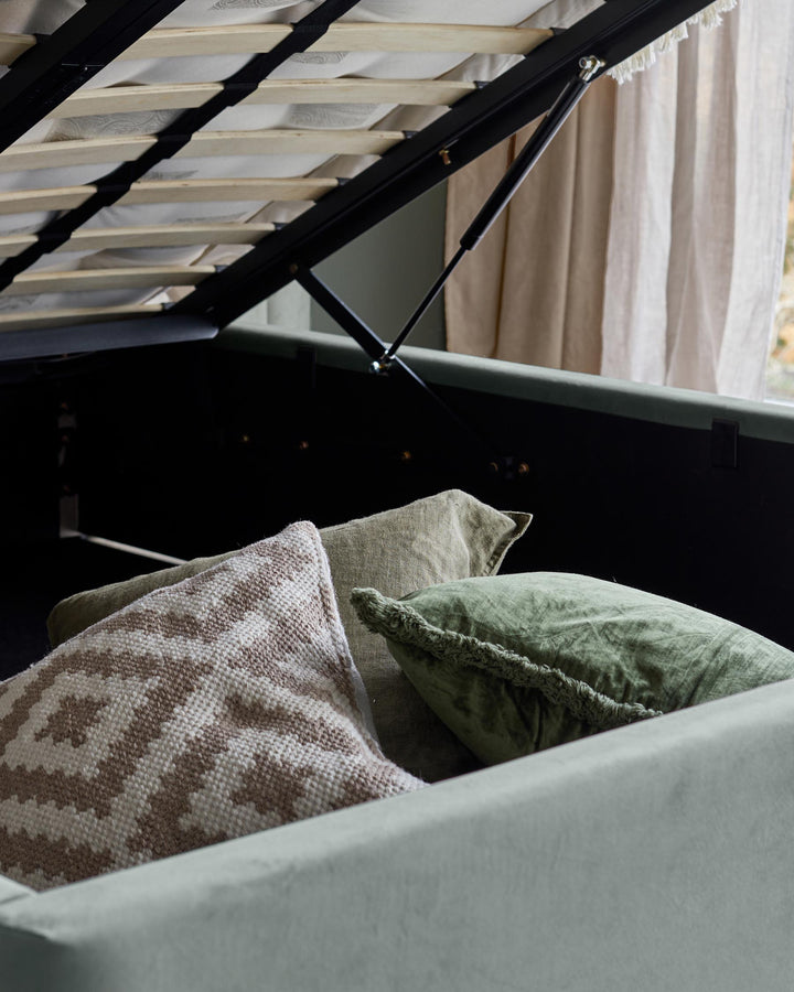 Upholstered ottoman bed with storage, cushions and a neutral curtain in the background